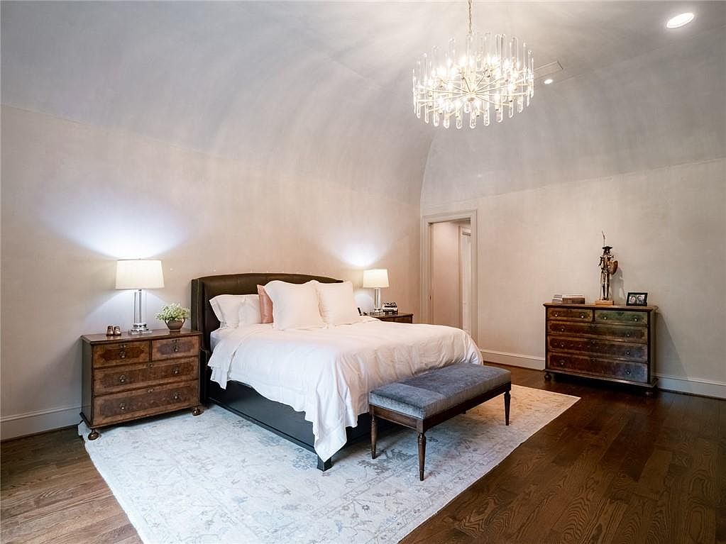 This spacious primary bedroom features a dramatic barrel-vaulted ceiling and a central crystal chandelier, creating an elegant and airy atmosphere. The room is anchored by a dark-framed bed with crisp white linens, flanked by two matching wooden dressers topped with table lamps. A plush grey bench sits at the foot of the bed, resting on a large, light-toned patterned rug that contrasts beautifully with the dark hardwood flooring.
