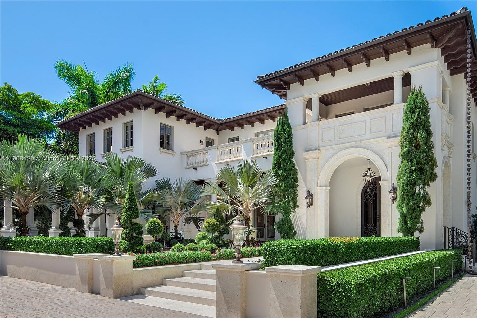 This is a front exterior view of a luxurious Mediterranean-style home. The house features a white facade, dark brown roof tiles, and an arched entryway. The landscaping is meticulously maintained with manicured hedges, palm trees, and decorative plants, creating a grand and inviting entrance.