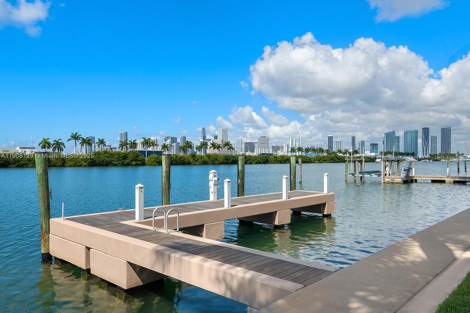 The image showcases a modern dock extending into a serene body of water, with a cityscape visible in the background. The dock features wooden planks and sturdy support structures, creating a clean and inviting space. The clear blue sky and calm water enhance the overall appeal, suggesting a peaceful and luxurious waterfront property.