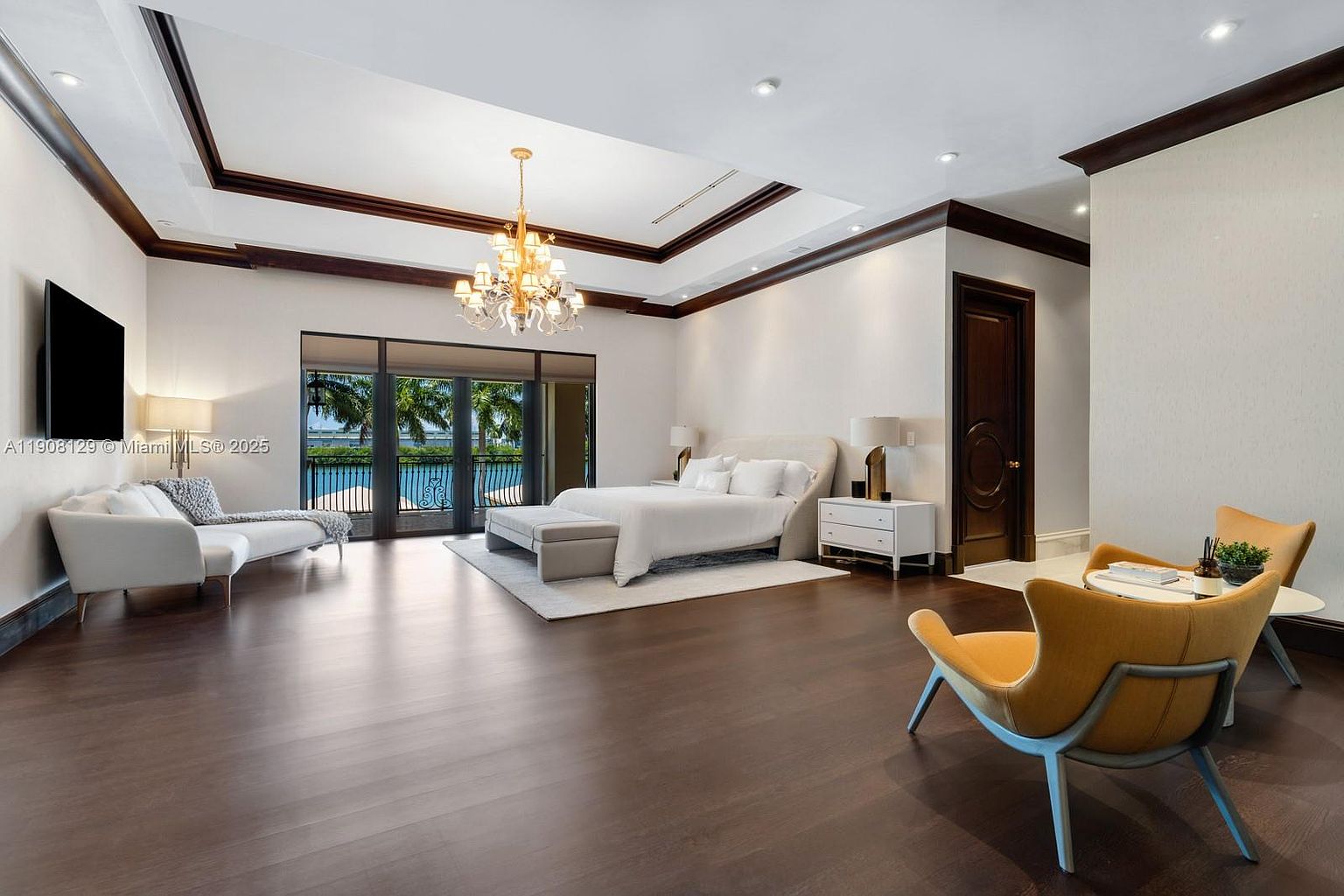 This is a spacious primary bedroom featuring dark hardwood floors, a tray ceiling with dark wood trim, and a large chandelier. The room includes a king-size bed with white linens, a seating area with a white chaise lounge, and a pair of mustard-colored chairs with a small table. Large sliding glass doors offer a view of the water, creating a luxurious and serene atmosphere.