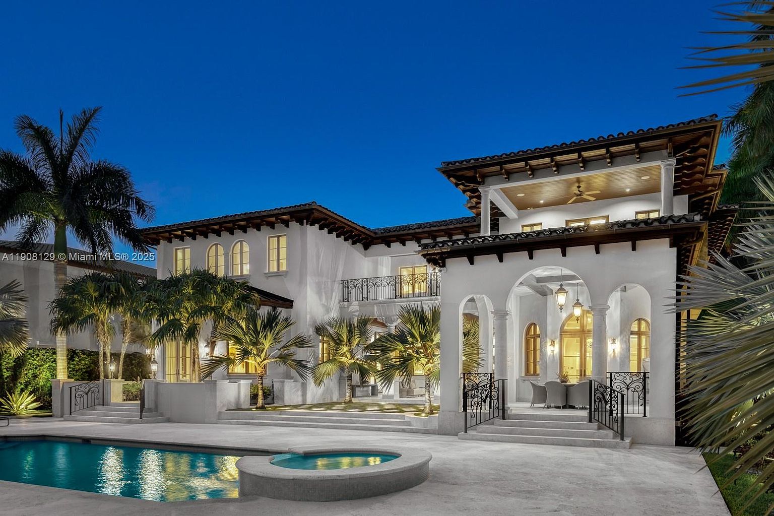 This is a rear view of a luxurious Mediterranean-style home at dusk, showcasing a pristine pool and spa area. The architecture features white stucco walls, dark tiled roofs, arched entryways, and wrought-iron balconies. Palm trees and manicured landscaping add to the property's appeal, creating a serene and upscale ambiance.