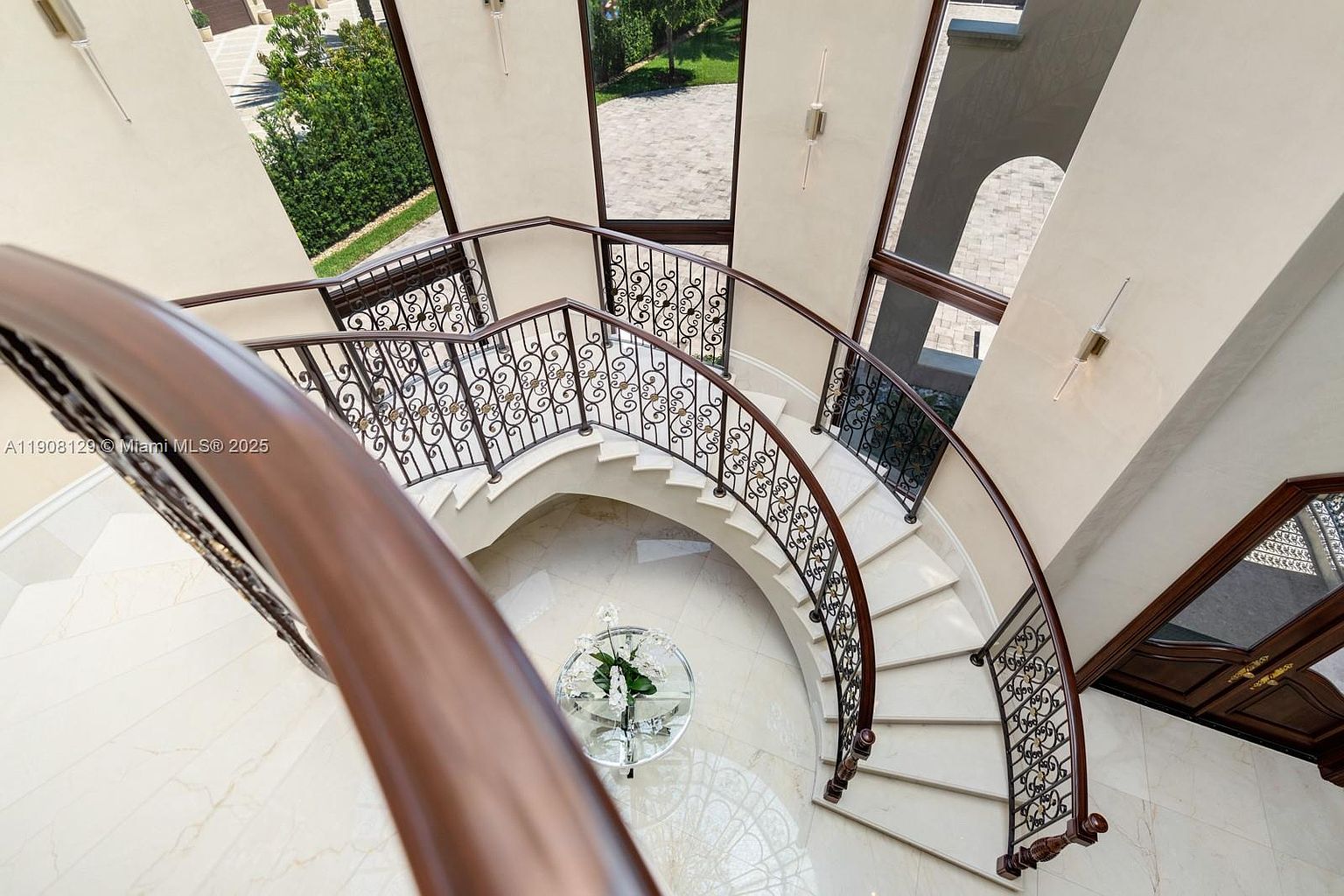 This is a high-angle shot of a grand, curved staircase inside a luxurious home. The staircase features white marble steps, ornate wrought-iron railings with a dark wood handrail, and a decorative glass table with floral arrangement at the base. Large windows provide natural light, highlighting the elegant architectural details and the spaciousness of the interior.