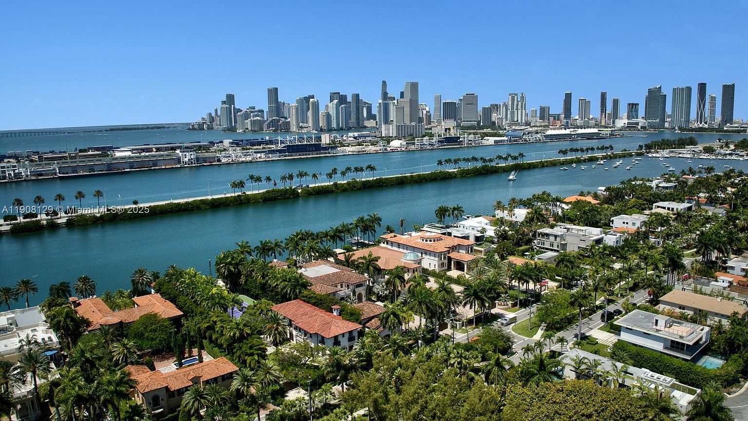 This aerial view showcases a luxurious waterfront neighborhood with stunning city skyline views. The scene features opulent homes with red-tiled roofs nestled among lush greenery and palm trees, bordering a tranquil waterway. The composition highlights the desirable location and upscale lifestyle associated with this real estate.