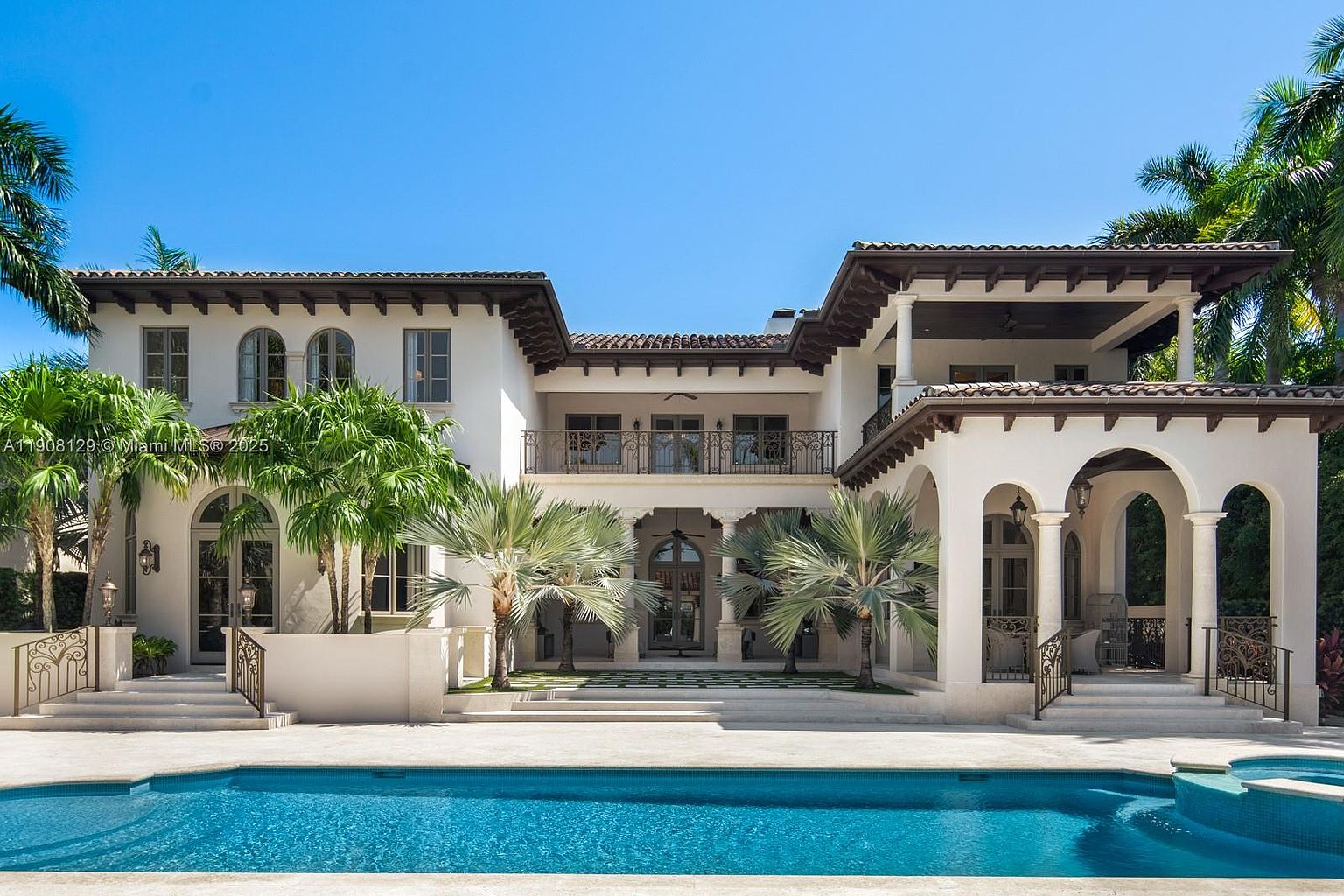 This image showcases the rear exterior of a luxurious Mediterranean-style home, emphasizing the pool and spa area. The architecture features white stucco walls, a red tile roof, arched openings, and wrought-iron balconies. Lush palm trees surround the property, enhancing the tropical ambiance and creating a serene outdoor living space.