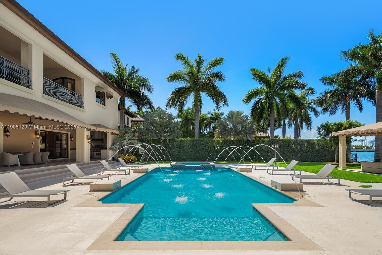 This image showcases a luxurious backyard pool area. The rectangular pool features fountains and is surrounded by lounge chairs and lush landscaping, including palm trees and manicured hedges. A covered seating area is visible in the background, suggesting a relaxing outdoor living space.