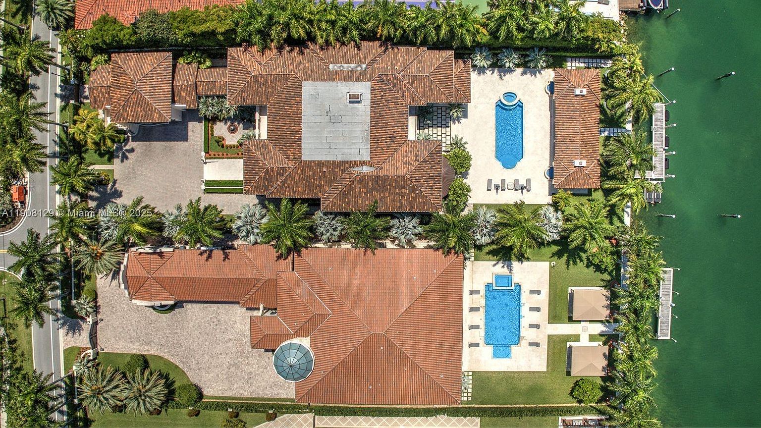 This aerial shot showcases two luxurious waterfront estates, each featuring terracotta-tiled roofs, private pools, and meticulously landscaped grounds with mature palm trees. The properties boast expansive layouts and direct access to the water, complete with docks, offering a glimpse into high-end coastal living. The perspective is from directly above, emphasizing the scale and exclusivity of the properties.