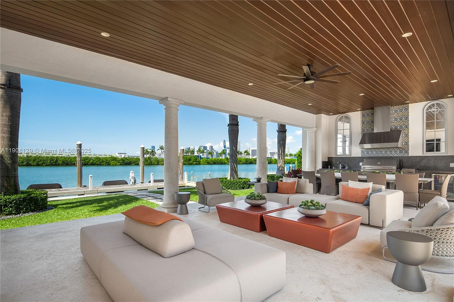 This image showcases a luxurious outdoor patio area with a stunning waterfront view. The space is furnished with modern, neutral-toned seating arrangements and stylish coffee tables, all under a covered area supported by elegant columns. An outdoor kitchen with stainless steel appliances is visible in the background, suggesting a perfect setting for entertaining and relaxation.