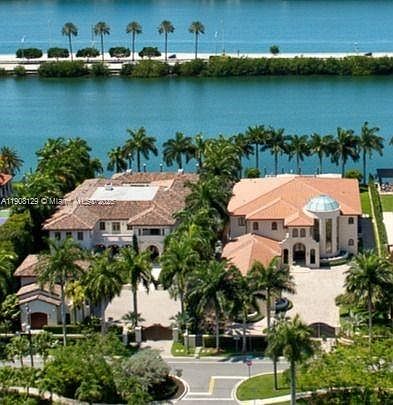 This aerial shot showcases a luxurious waterfront property with meticulously landscaped grounds. The residence features a terracotta tile roof, a prominent rotunda, and a sprawling driveway. The property is surrounded by lush greenery and offers stunning water views, creating an impression of exclusivity and opulence.