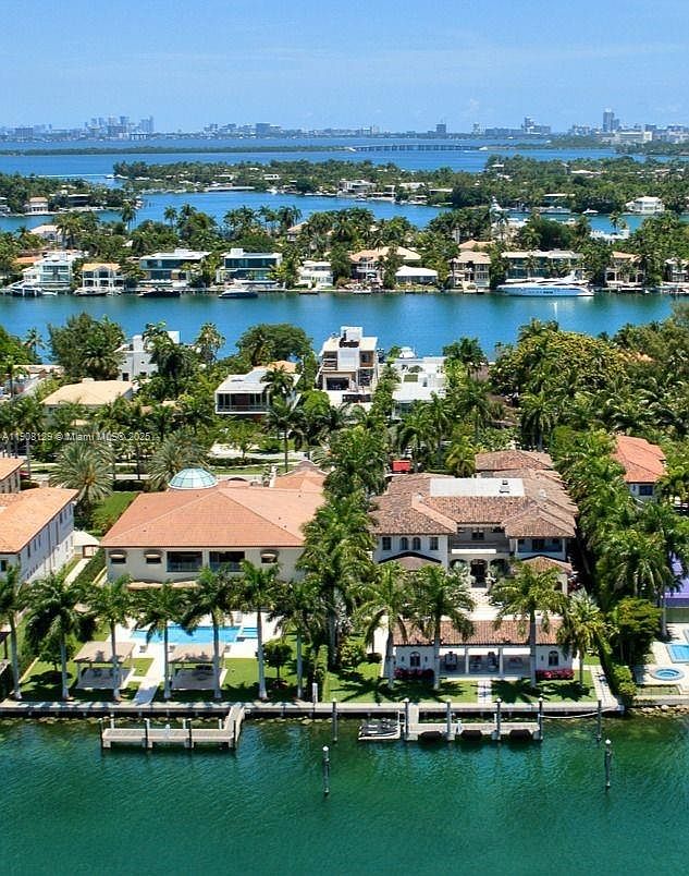 This aerial view showcases luxurious waterfront properties with lush landscaping and private docks. The homes feature Mediterranean-style architecture with red tile roofs and multiple outdoor living spaces. The turquoise water and distant city skyline create a stunning backdrop, emphasizing the exclusivity and high-end nature of the real estate.