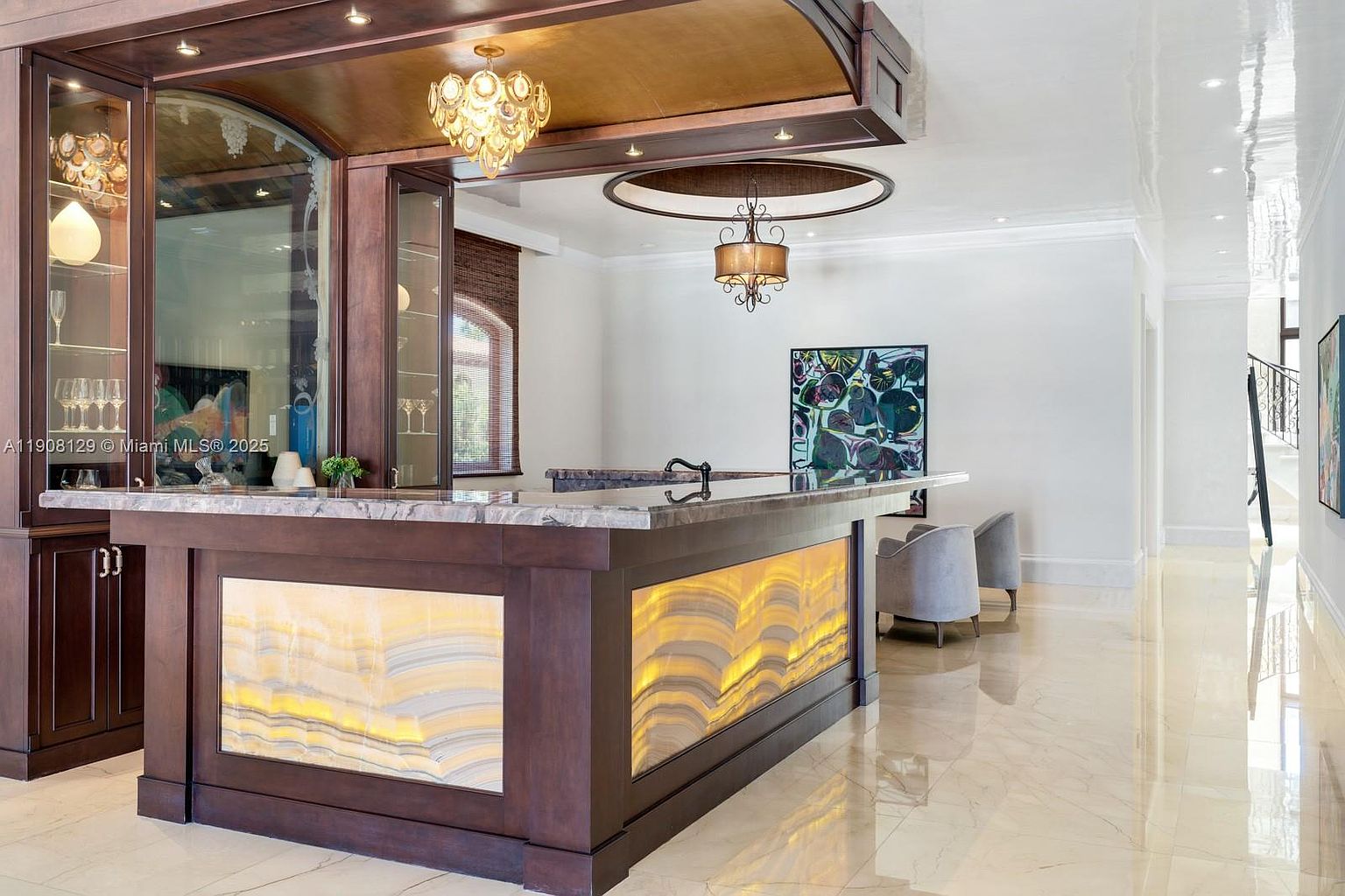 This interior shot showcases a luxurious bar area within a living space, featuring a marble countertop, illuminated onyx panels, and custom cabinetry. The room is well-lit with decorative chandeliers and recessed lighting, complemented by polished marble flooring that reflects the light, creating an elegant and inviting atmosphere.