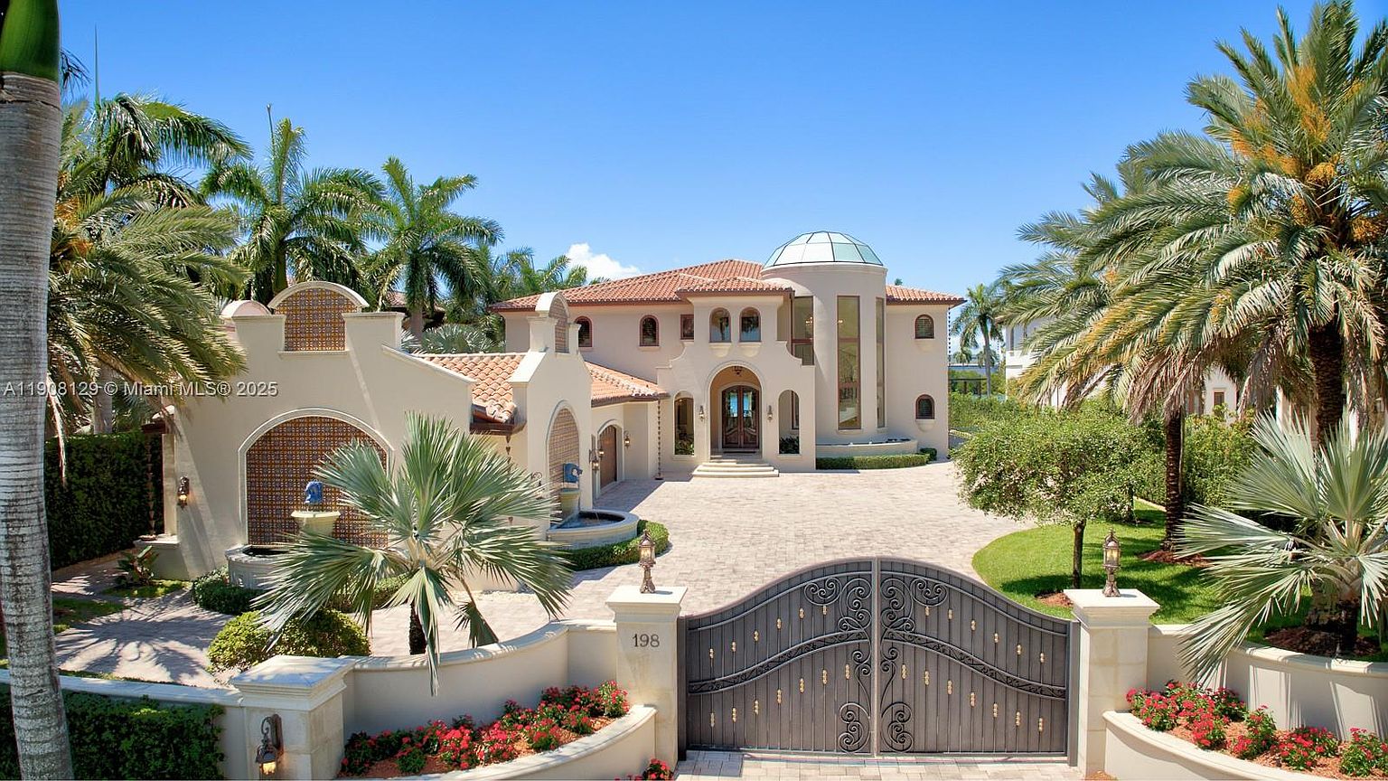 This is a front exterior view of a luxurious Mediterranean-style home. The property features a grand entrance gate, a circular driveway, a terracotta tile roof, and lush tropical landscaping with palm trees. The house has a light-colored stucco exterior, arched windows, and a distinctive glass-domed tower, creating an impression of elegance and sophistication.