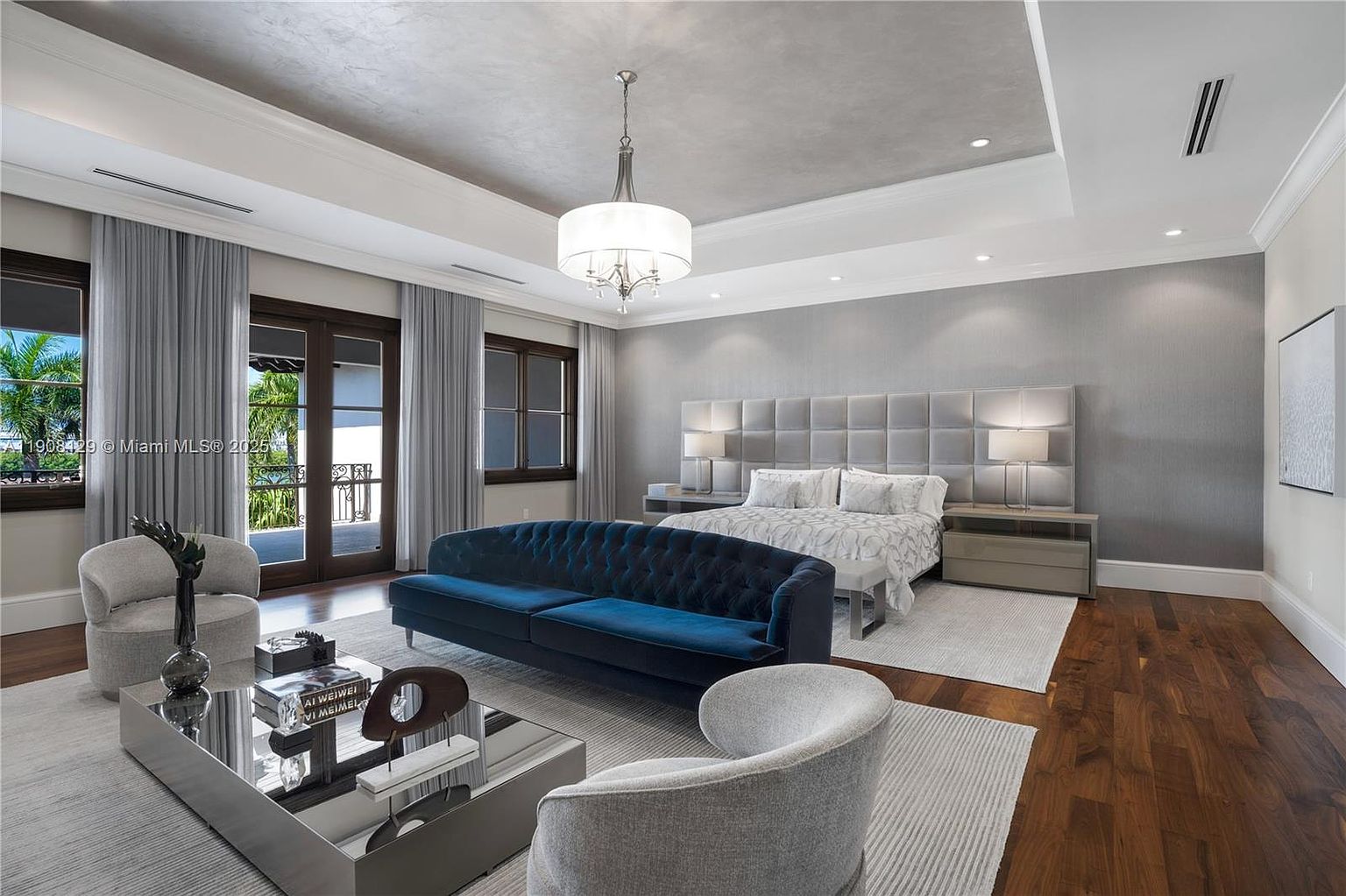 This is a luxurious primary bedroom featuring a large bed with a tufted headboard, flanked by matching nightstands and lamps. A stylish blue velvet sofa sits in the foreground, complemented by modern armchairs and a reflective coffee table. The room is well-lit with a chandelier and recessed lighting, and the hardwood floors add warmth to the space.