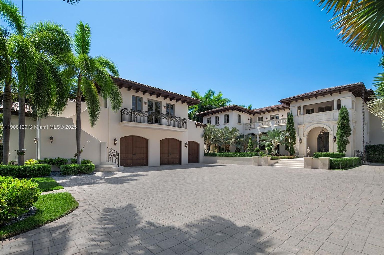 This is a front exterior view of a luxurious Mediterranean-style home. The property features a large paved driveway, multiple garage doors, a balcony with wrought iron railings, and meticulously manicured landscaping with palm trees and hedges. The architecture showcases a light-colored facade with a dark tile roof, creating an elegant and inviting curb appeal.