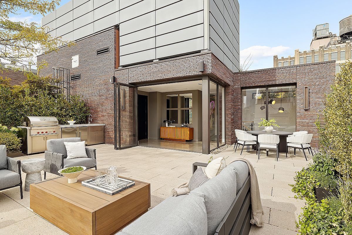 This image showcases a luxurious outdoor patio area, featuring modern furniture such as a gray sofa, armchairs, and a wooden coffee table. An outdoor kitchen with a stainless steel grill and sink is visible, along with a dining area set with a table and chairs. The patio is paved with light-colored stone, and the surrounding architecture includes brick and metal cladding, creating a sophisticated and inviting outdoor living space.