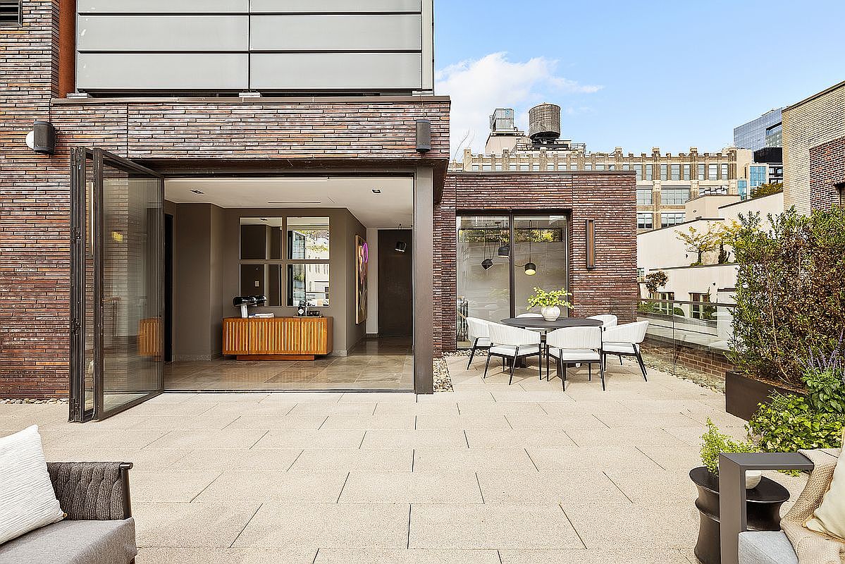 This image showcases a luxurious outdoor patio area, seamlessly blending indoor and outdoor living. The space features a dining set with modern chairs, a well-maintained stone patio, and a partial view of the interior through large, open doors. The surrounding architecture includes brickwork and glimpses of the cityscape, adding to the upscale urban feel.