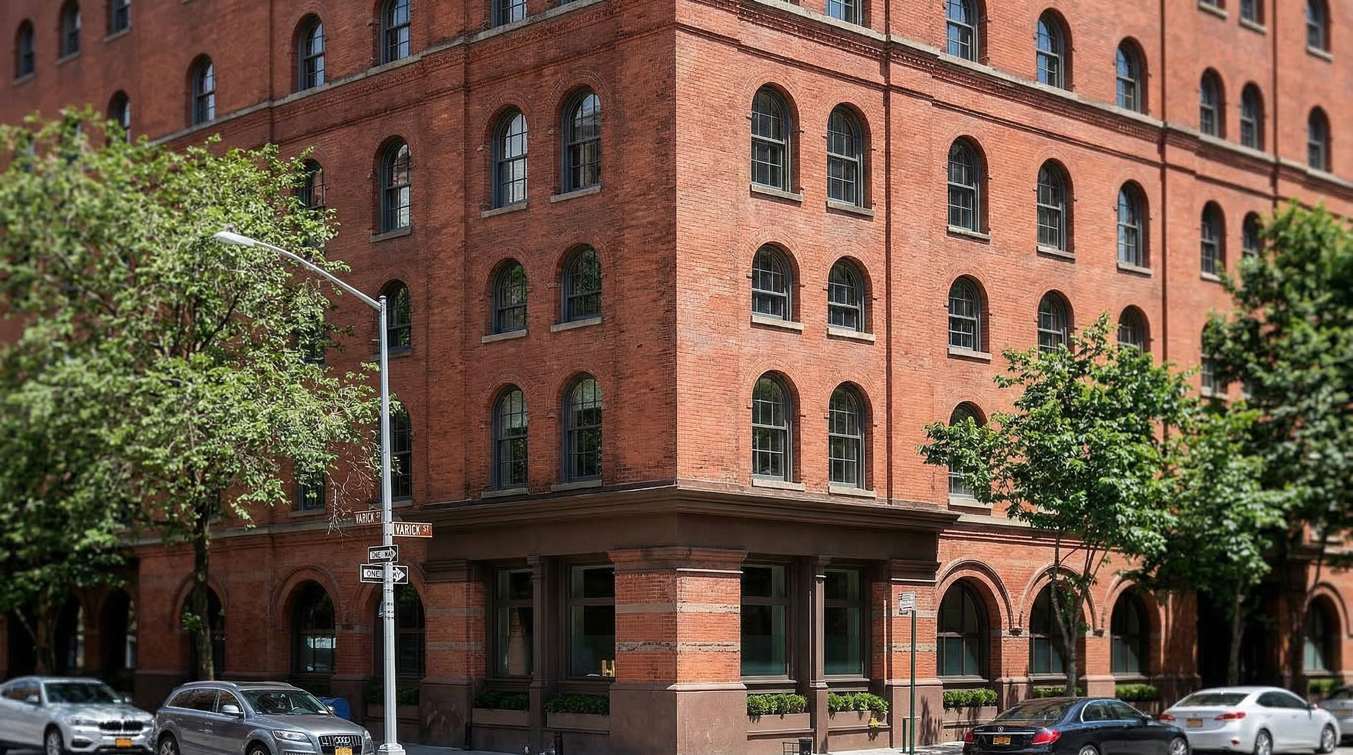 This building exterior showcases a classic, historic industrial-style architecture characterized by its distinct red-brick facade and arched window detailing. The structure occupies a prominent corner lot location, adding to its urban appeal and presence. The presence of mature trees and urban street identifiers highlights its convenient setting within a vibrant neighborhood.
