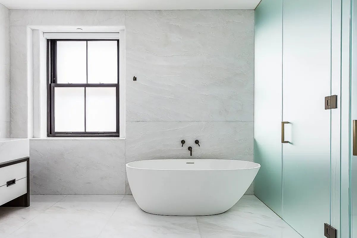 This is a modern primary bathroom featuring a freestanding white bathtub with wall-mounted fixtures. The walls are covered in large-format marble-like tiles, and a black-framed window provides natural light. A frosted glass shower enclosure with bronze hardware is visible to the right, creating a spa-like atmosphere.