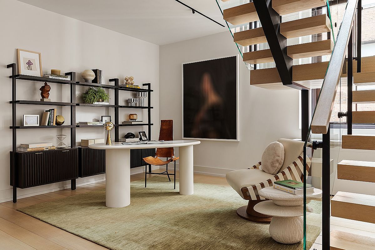 This interior shot showcases a stylish home office featuring a modern aesthetic. A black shelving unit displays various decorative items and books, complementing the sleek white desk and a comfortable chair. The room is well-lit and includes a unique piece of art, creating an inviting and productive workspace, with a staircase in the background.