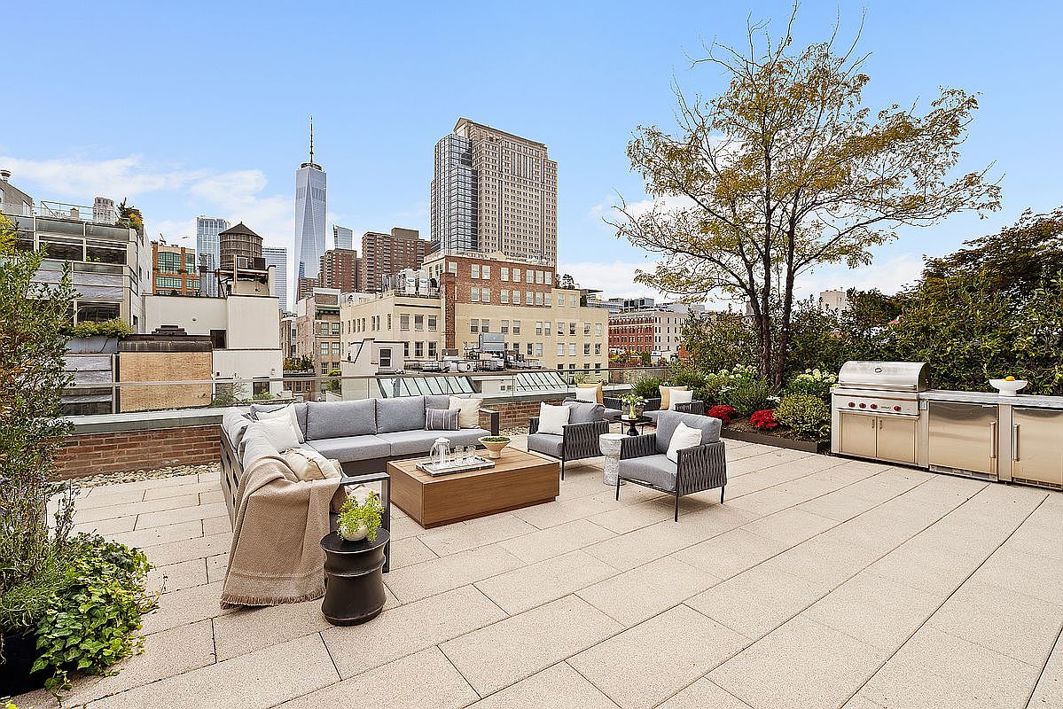 This is a spacious rooftop patio featuring comfortable outdoor seating, including a gray sofa, armchairs, and a wooden coffee table. A stainless steel outdoor kitchen with a grill is visible in the background, surrounded by greenery and flowers. The patio offers stunning city views, including the One World Trade Center, creating a luxurious and inviting outdoor living space.