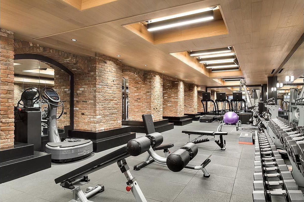 This is a well-equipped home gym featuring brick archways and modern lighting. The space includes various exercise machines, free weights, and a vibration plate. The flooring is a dark tile, and the overall impression is a high-end, dedicated fitness area.