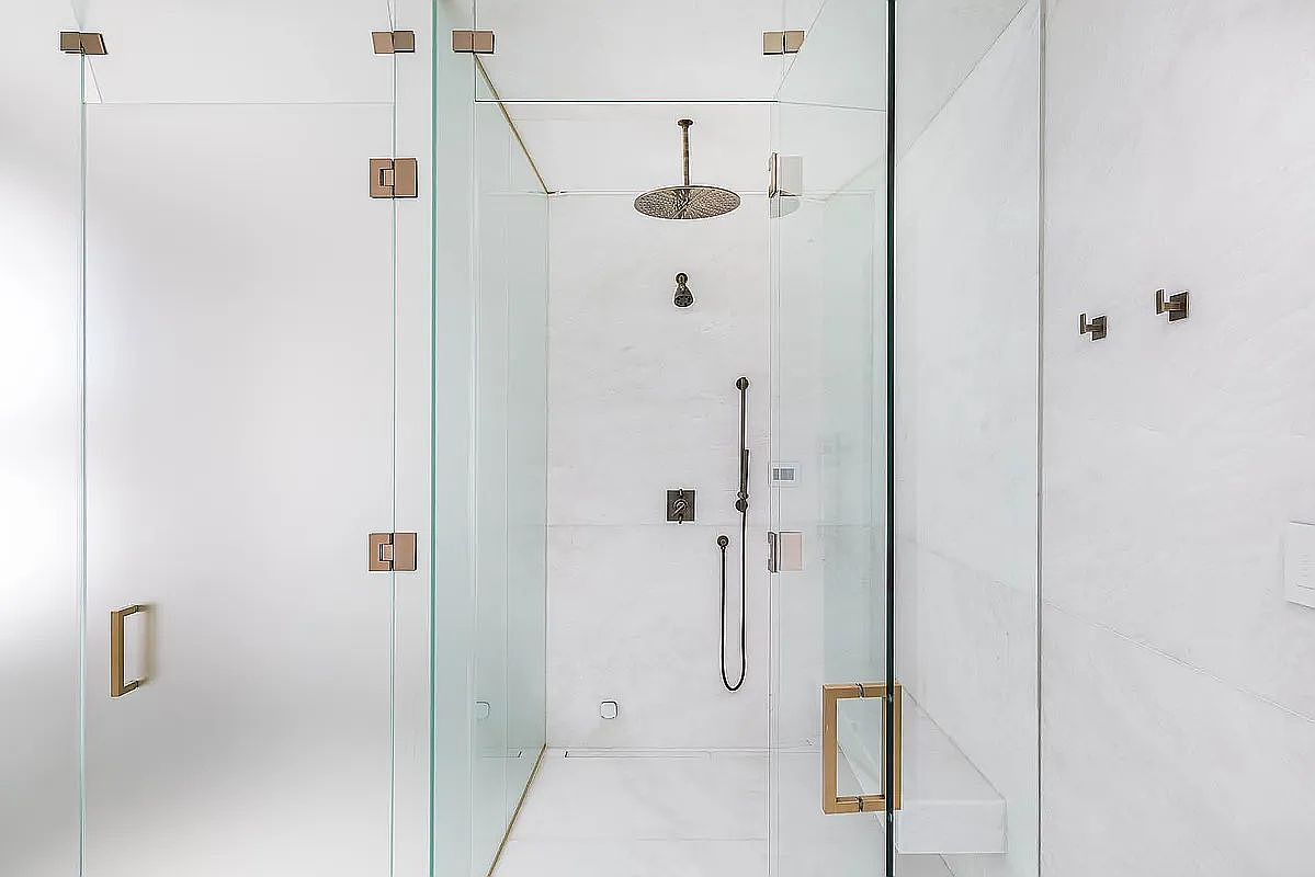 This is a modern primary bathroom featuring a glass-enclosed shower with bronze hardware. The shower has a rainfall showerhead and a handheld sprayer. The walls are white marble, and there is a built-in bench. The overall style is minimalist and luxurious.