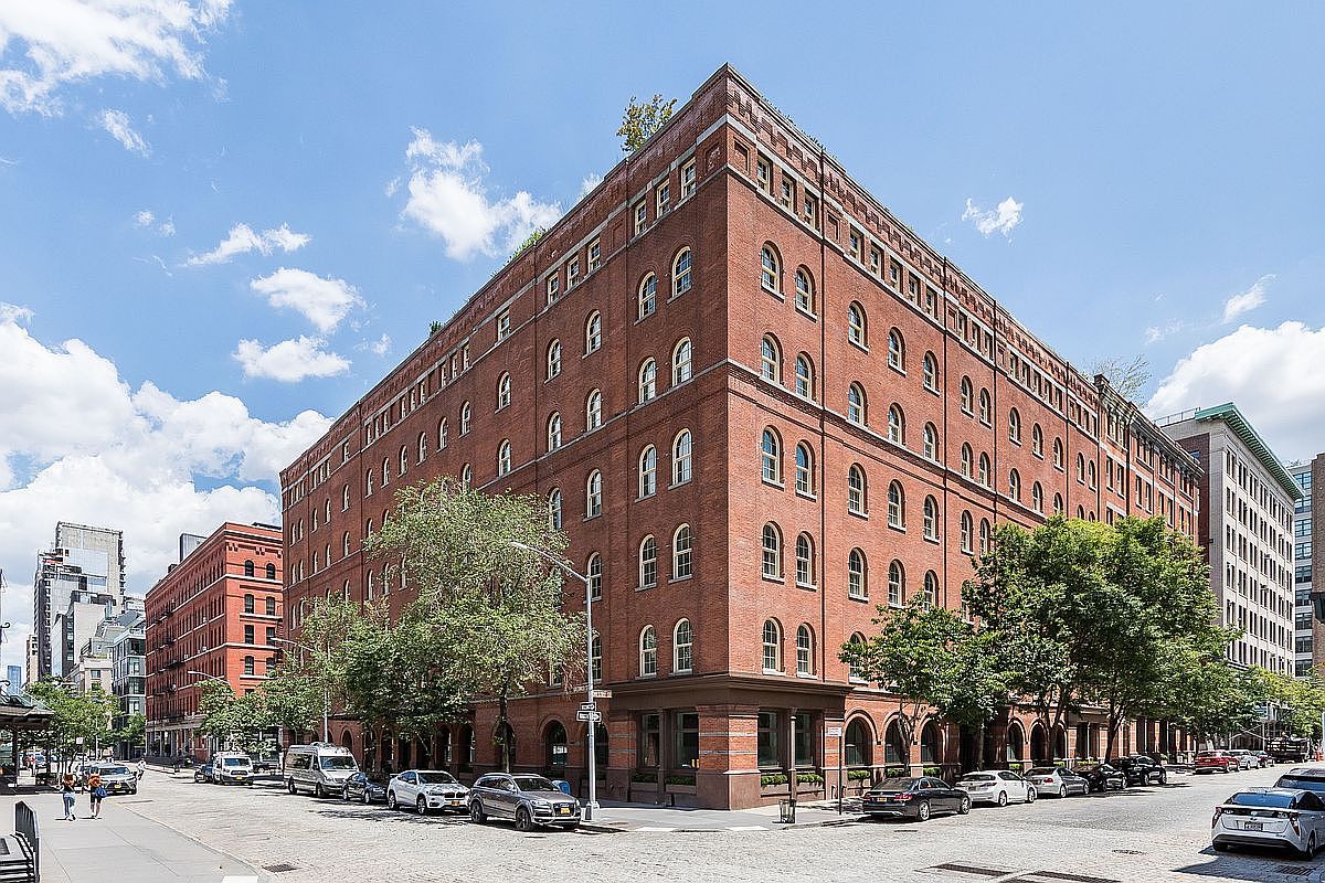 This is an exterior shot of a large, red brick building with arched windows, showcasing its impressive size and architectural details. The building occupies a corner lot, with street views and mature trees adding to the urban landscape. Several cars are parked along the street, and pedestrians are visible in the background, giving a sense of the building's location within a vibrant city environment.