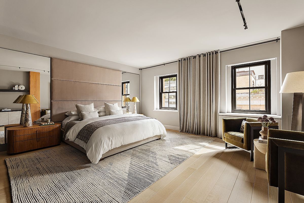 This is a spacious primary bedroom featuring a large bed with a padded headboard, neutral-toned bedding, and a decorative throw. The room is well-lit with natural light from two windows dressed with elegant curtains. The flooring is light hardwood, and a patterned rug adds texture to the space, creating a serene and inviting atmosphere.