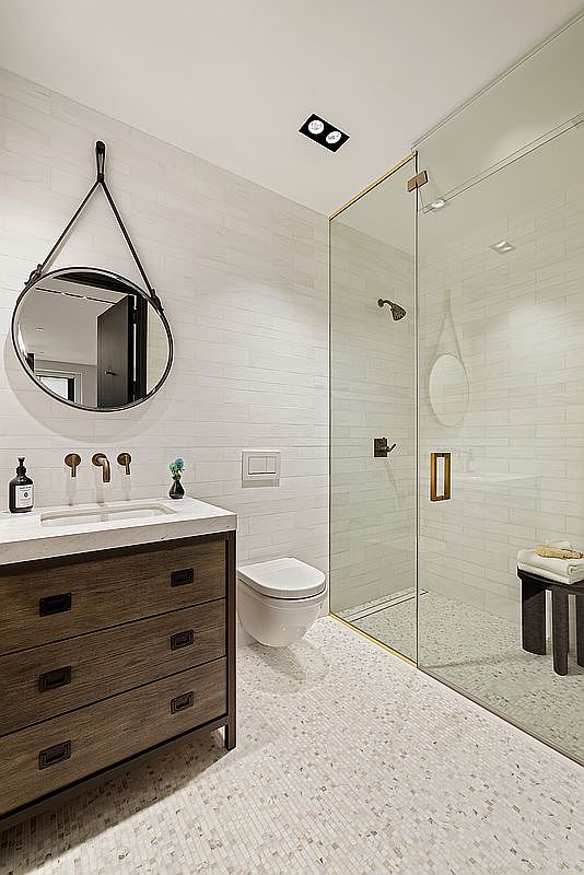 This is a well-lit primary bathroom featuring a modern design with white subway tile walls and mosaic tile flooring. A wooden vanity with a light countertop and a round mirror add warmth to the space. A glass-enclosed shower with matching tiles completes the luxurious feel of the bathroom.