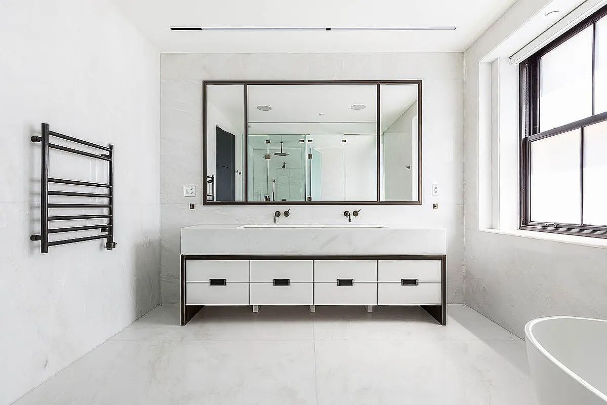 This is a bright and modern primary bathroom featuring a double vanity with a white countertop and sleek cabinetry. A large, framed mirror hangs above the vanity, reflecting a glimpse of a glass-enclosed shower. A black towel warmer is mounted on the wall, and a large window provides natural light, enhancing the clean and minimalist aesthetic.