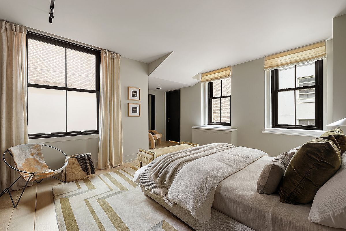 This primary bedroom features a neutral color palette with a large bed adorned with plush pillows and a cozy blanket. Natural light streams in through multiple windows, some with roman shades, enhancing the room's serene atmosphere. A stylish chair and a patterned rug add subtle visual interest, creating a comfortable and inviting space.