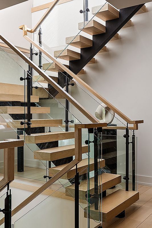 This image showcases a modern staircase with floating wooden treads and glass railings, creating an open and airy feel. Black metal supports add a contemporary touch, while the light wood finish complements the minimalist design. The staircase is well-lit and appears to be a central architectural feature of the home.