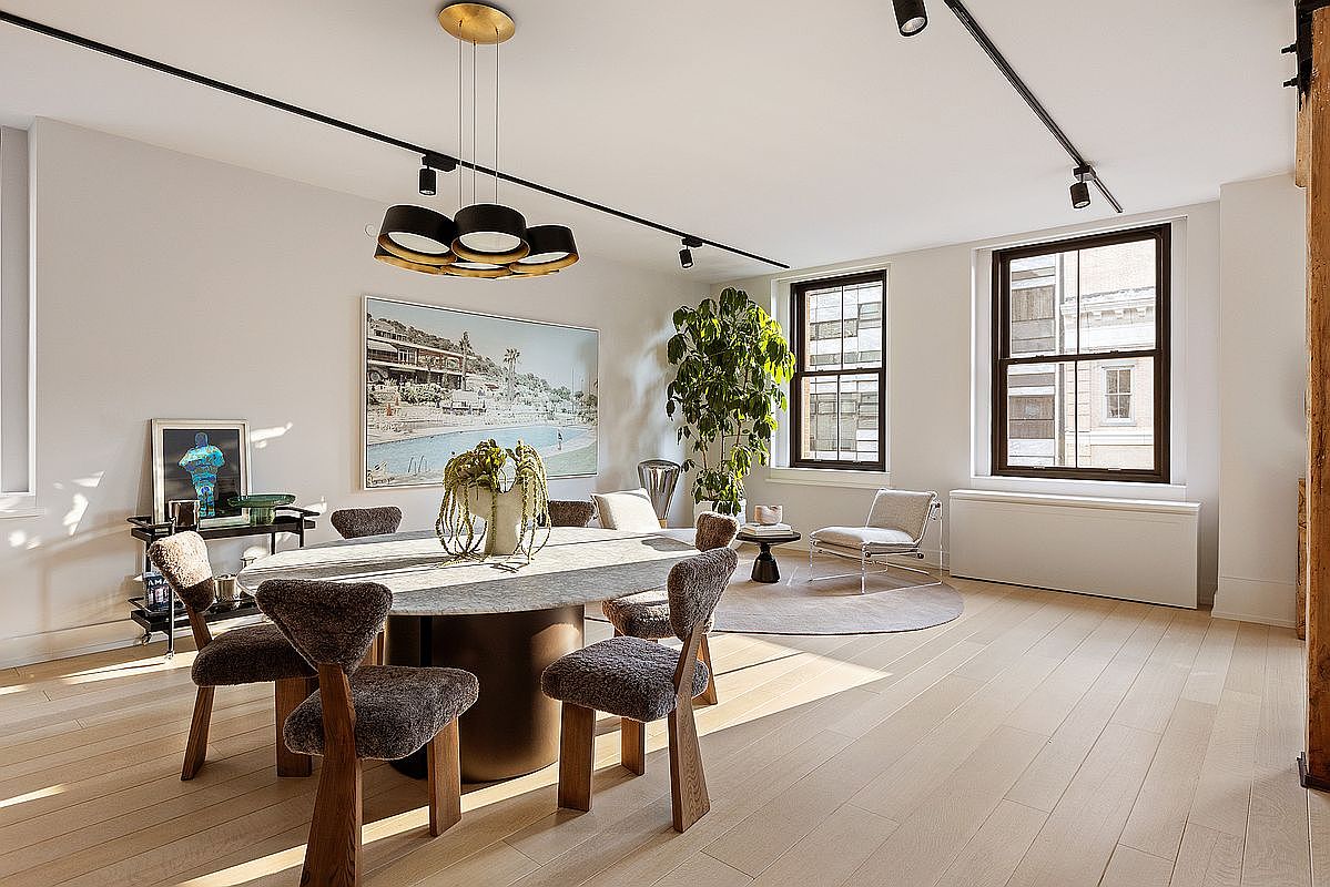 This is an interior shot of a dining room featuring a large oval table with unique, upholstered chairs. A modern chandelier hangs above the table, and artwork adorns the walls. Natural light streams in through the windows, highlighting the hardwood floors and creating a bright and inviting atmosphere.