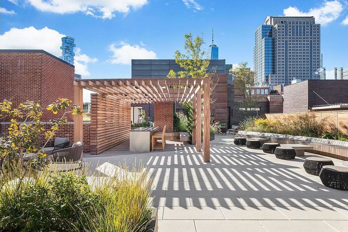 This image showcases a stylish rooftop patio/deck area with a modern pergola, outdoor seating, and lush greenery. The space features a built-in outdoor kitchen area and comfortable lounge seating, creating an inviting atmosphere for relaxation and entertainment. The backdrop includes city skyline views, adding to the property's appeal.