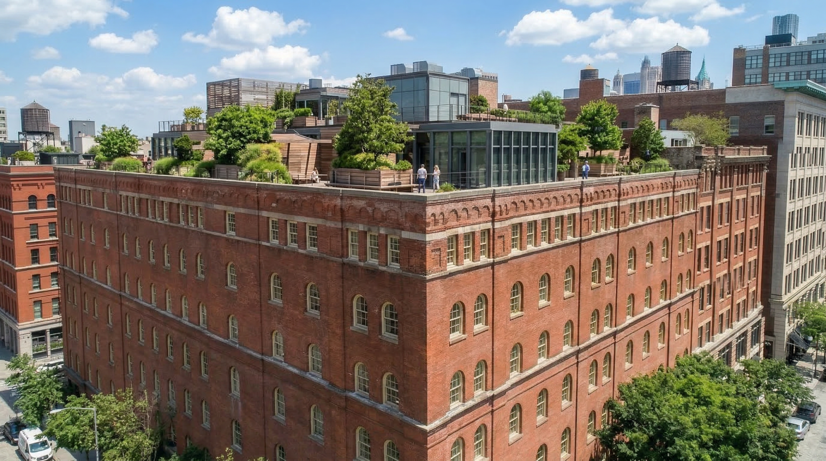 This stunning high-angle exterior view highlights a historic red-brick loft building featuring a meticulously landscaped rooftop terrace. The terrace includes mature plantings, wooden decking, and modern glass-enclosed structures, seamlessly blending industrial architecture with luxury outdoor living. The scene captures an expansive urban neighborhood setting, reflecting a premium real estate conversion typical of highly desirable city centers.
