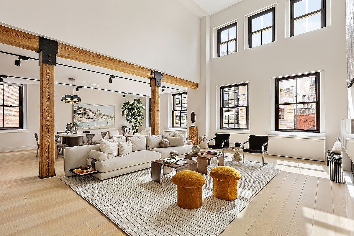 This is a spacious and modern living room featuring high ceilings and large windows that provide ample natural light. The room is furnished with a large sectional sofa, wooden coffee tables, and unique mushroom-shaped ottomans, all set on a striped area rug. Exposed wooden beams add a touch of rustic charm to the contemporary design.