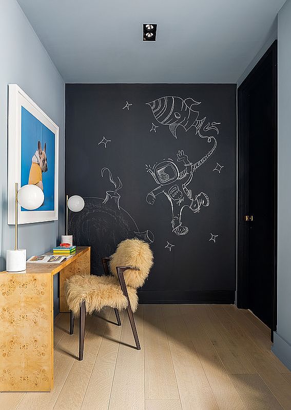 This interior shot showcases a stylish and creative office space. A light wood desk with a unique burl wood pattern is paired with a furry chair, set against a chalkboard wall featuring whimsical space-themed drawings. The room is illuminated by modern globe lamps, creating a playful yet sophisticated atmosphere.