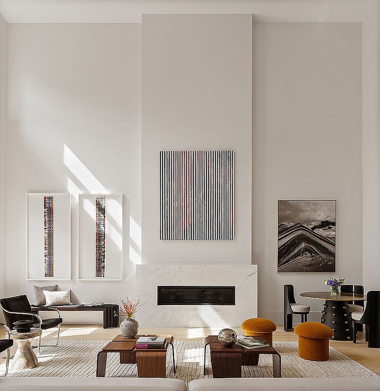 This is a spacious living room with high ceilings and a modern aesthetic. The room features a fireplace with a marble surround, contemporary artwork, and stylish furniture including a round dining table with chairs and a pair of orange ottomans. Natural light floods the space, creating a bright and inviting atmosphere.