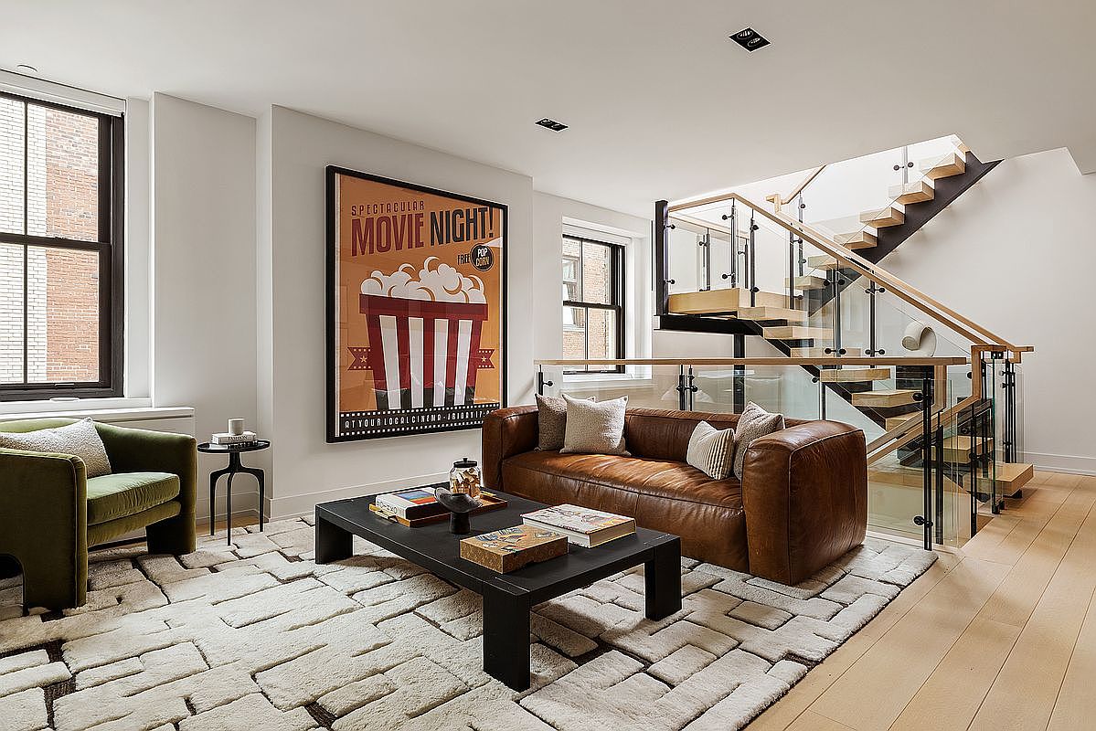 This is a stylish living room featuring a brown leather sofa, a black coffee table with books, and a large movie poster on the wall. The room has a modern staircase with glass railings and a textured rug that adds warmth. The space is well-lit with natural light from the windows, creating a cozy and inviting atmosphere.
