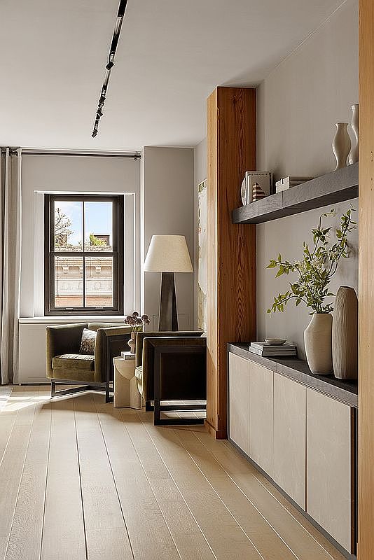 This interior shot showcases a stylish living room with hardwood floors and neutral-toned walls. The room features modern furniture, including a pair of armchairs, a dark wood side table, and a built-in shelving unit with decorative items. Natural light streams in through a window, creating a warm and inviting atmosphere.