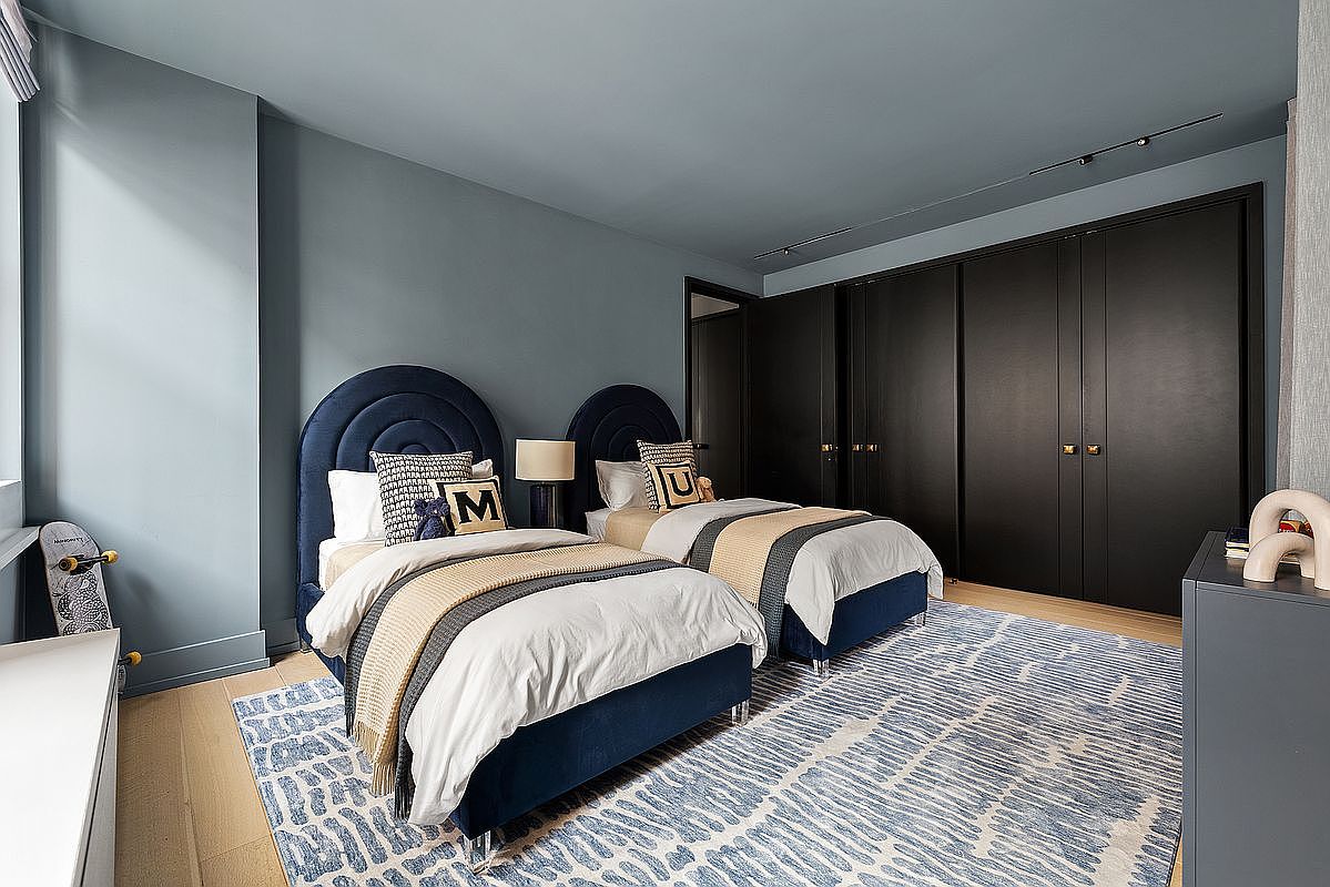 This is a well-designed guest bedroom featuring two twin beds with navy blue upholstered headboards and matching bed frames. The room is decorated in a modern style with a blue and white patterned rug, blue walls, and dark closets. The room feels cozy and inviting, perfect for a real estate listing showcasing a home's versatility.