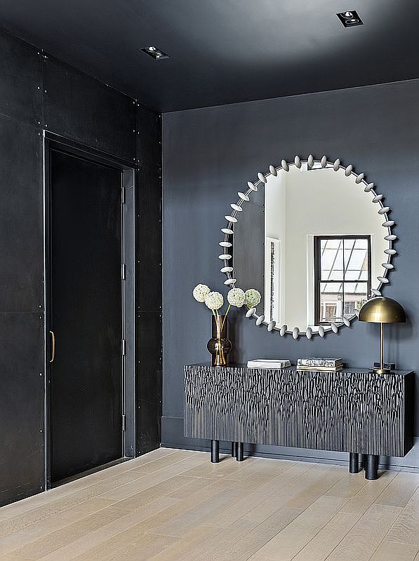 This interior shot showcases a modern hallway with dark walls and a light wood floor. A black door is visible on the left, while a decorative console table with a round mirror and a small lamp adorns the right wall. The space exudes a sophisticated and contemporary aesthetic, perfect for a stylish home.