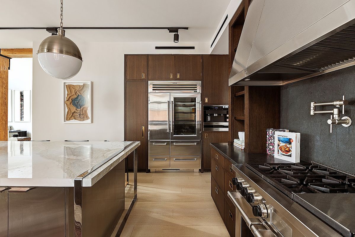 This is a well-appointed kitchen featuring dark wood cabinetry and stainless steel appliances, including a large refrigerator and professional-grade range. A marble-topped island provides ample counter space, and modern lighting fixtures illuminate the area. The overall impression is one of luxury and functionality.