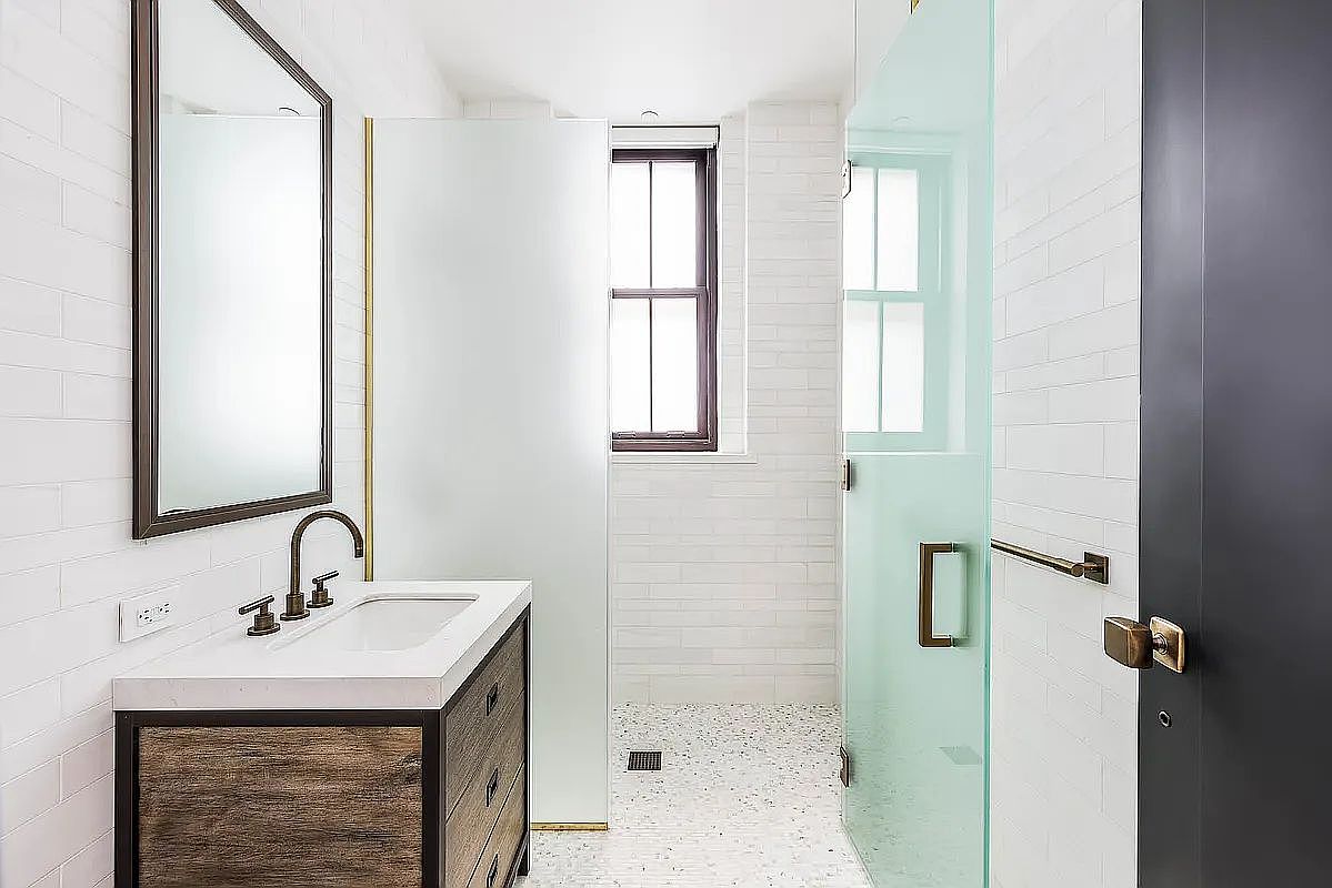 This is a well-lit bathroom featuring a modern vanity with a white countertop and a rustic wood-paneled base. A rectangular mirror hangs above the sink, and the walls are tiled with white subway tiles. The shower area is enclosed with a frosted glass panel and a clear glass door, and the floor is covered in small, speckled tiles, creating a clean and contemporary aesthetic.