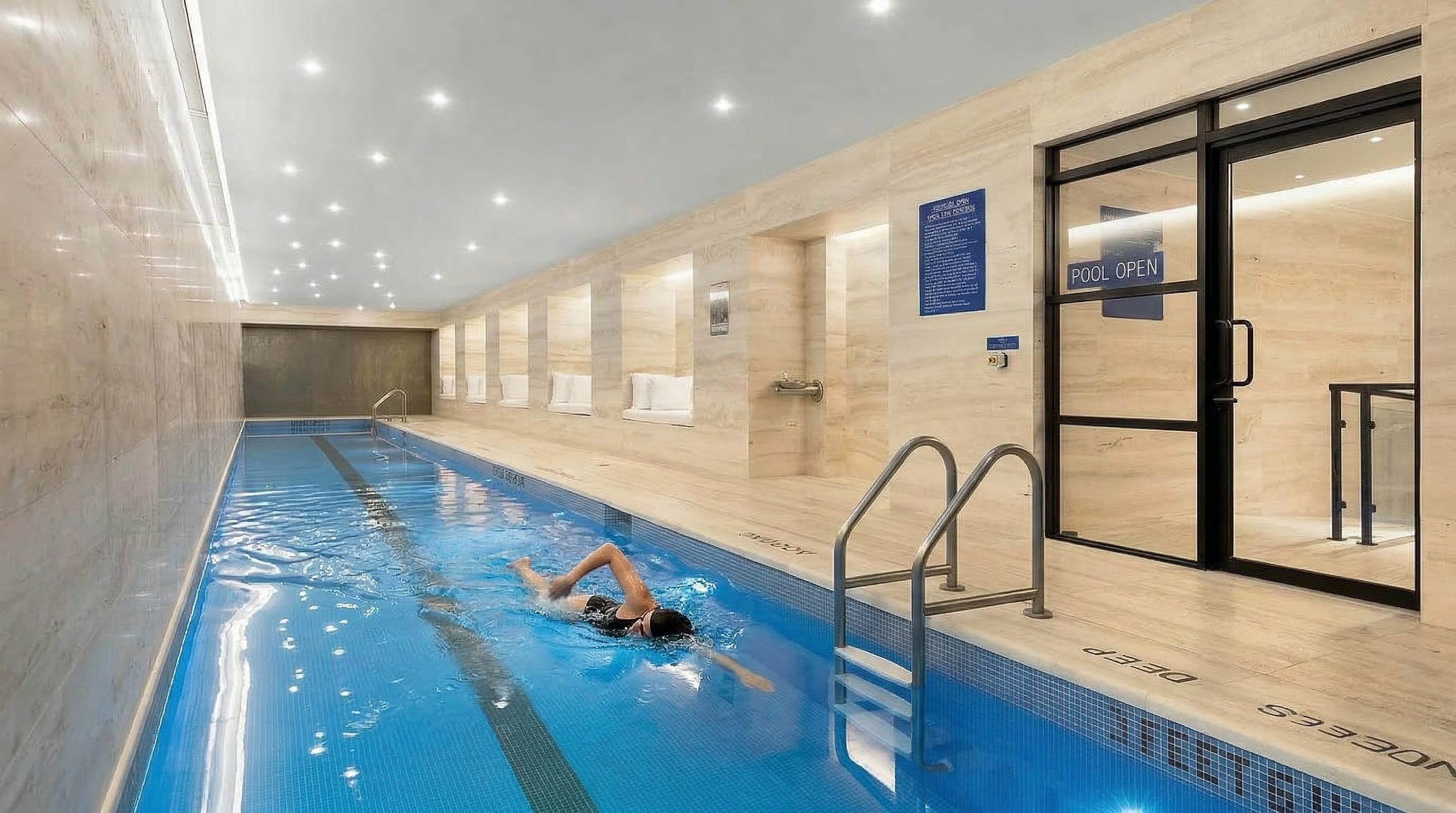 This image showcases an indoor swimming pool area, featuring a long, rectangular pool with blue mosaic tiles and a dark stripe down the center. The pool is surrounded by light-colored stone or tile, with recessed seating areas along one side and a glass door leading to another area. The space is well-lit with recessed lighting, creating a clean and luxurious atmosphere.