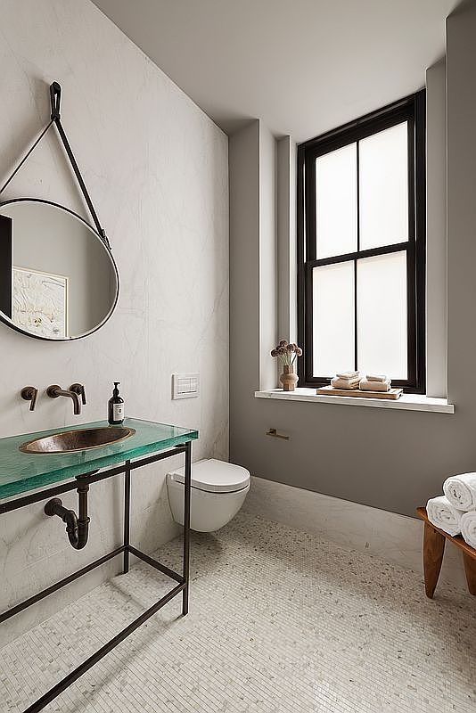 This is a stylish bathroom featuring a unique glass-topped vanity with a copper sink and bronze fixtures. A round mirror hangs above the sink, complemented by a wall-mounted toilet and a window providing natural light. The floor is tiled with small mosaic tiles, adding texture to the space.