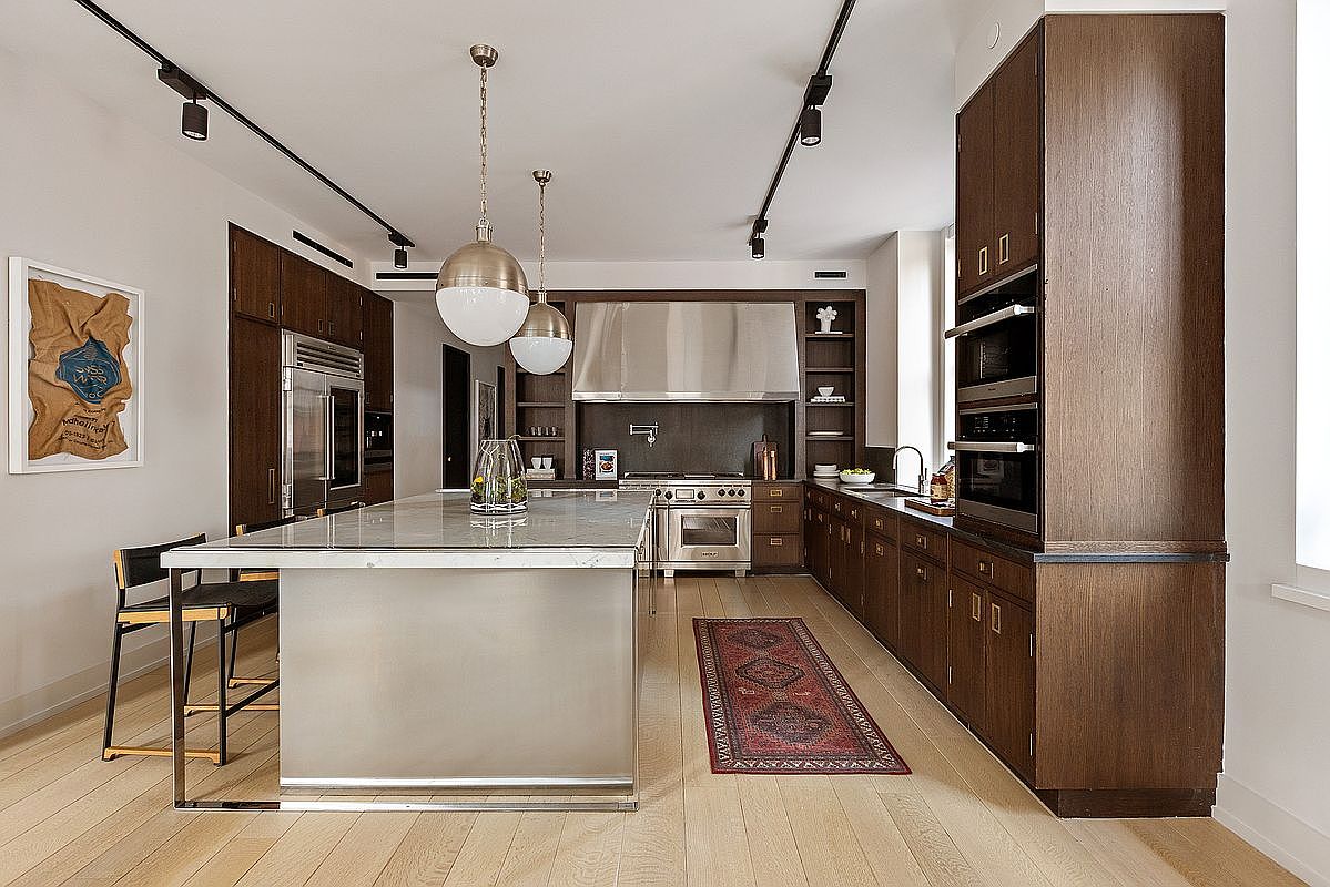 This is a well-lit kitchen featuring dark wood cabinetry, stainless steel appliances, and a large island with a marble countertop. The kitchen has a modern design with track lighting and pendant lights. A red patterned rug adds a touch of color to the light wood flooring.