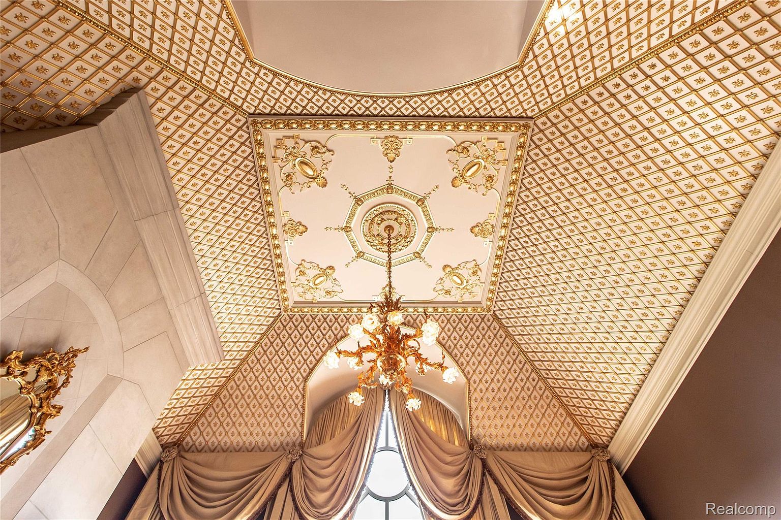 This image showcases an opulent interior architectural detail, focusing on the ceiling and a grand chandelier. The ceiling features intricate gold detailing and patterns, creating a luxurious and elegant atmosphere. A large window with draped curtains adds to the grandeur, suggesting a high-end property with exquisite design elements.