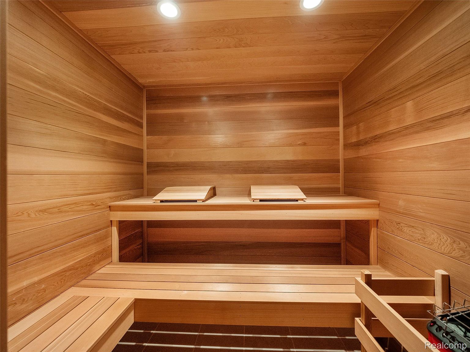 This is an interior shot of a sauna, featuring wooden walls, benches, and flooring. The sauna is well-lit with overhead lights, creating a warm and inviting atmosphere. The clean lines and natural wood give it a minimalist and relaxing aesthetic.