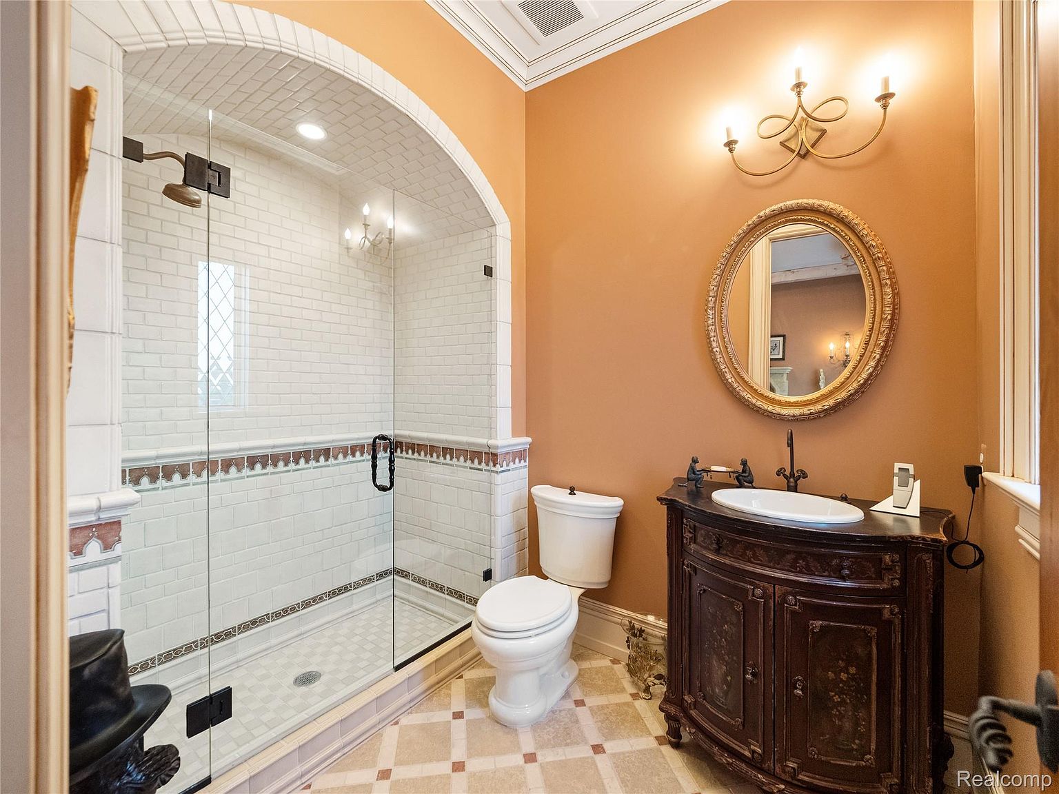This is a well-appointed primary bathroom featuring a glass-enclosed shower with white subway tile and decorative trim, a toilet, and a dark wood vanity with an oval sink and ornate gold-framed mirror. The walls are painted a warm orange hue, and the flooring is a patterned tile. The overall impression is luxurious and classic.
