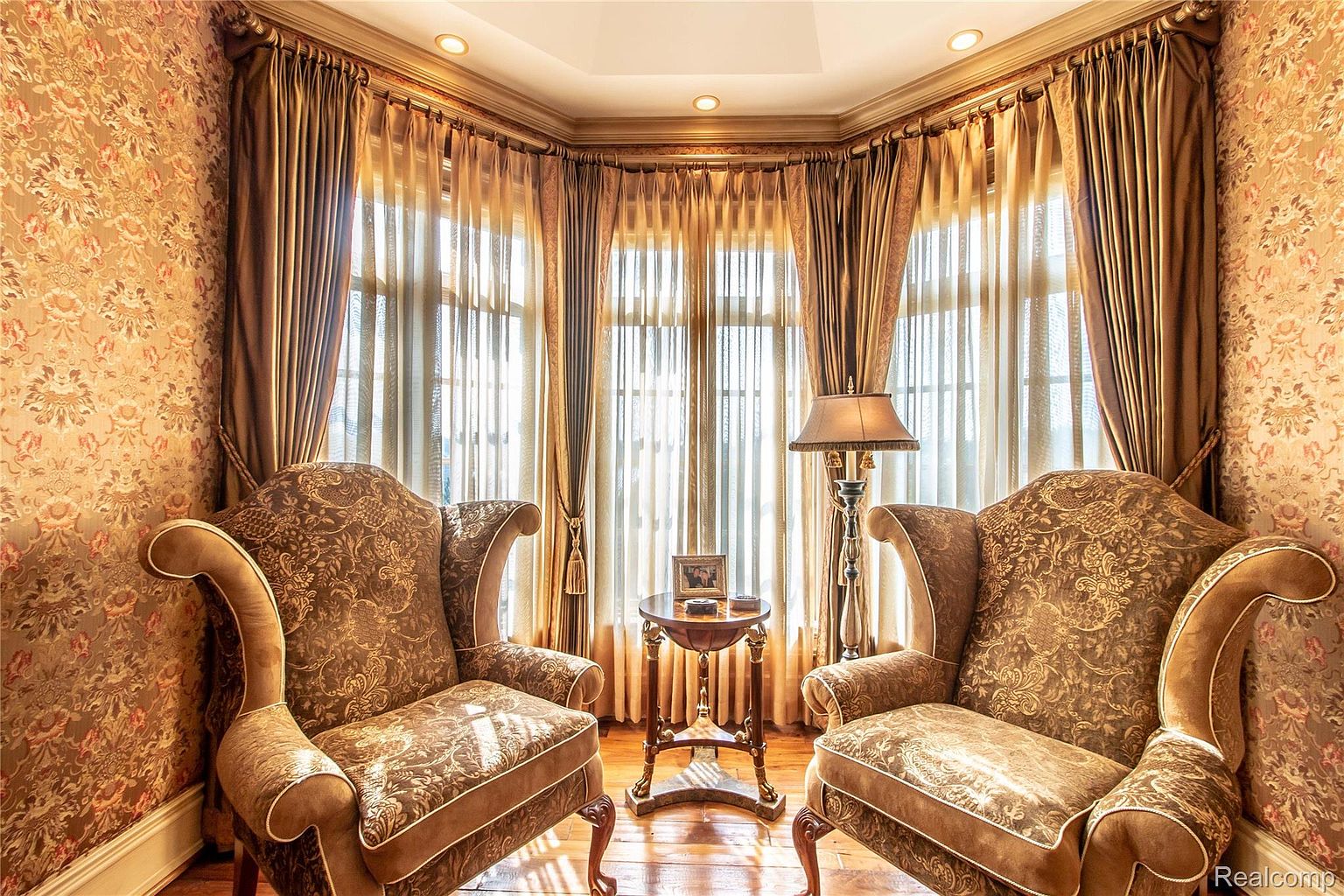 This interior shot showcases a cozy living room with two ornate armchairs flanking a small table with a lamp. The room is bathed in warm light filtering through sheer curtains, complemented by rich brown drapes. The walls are adorned with patterned wallpaper, creating a traditional and inviting atmosphere.